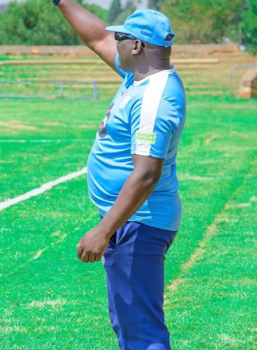 Coach in a light blue football kit and cap gesturing at the side of a pitch